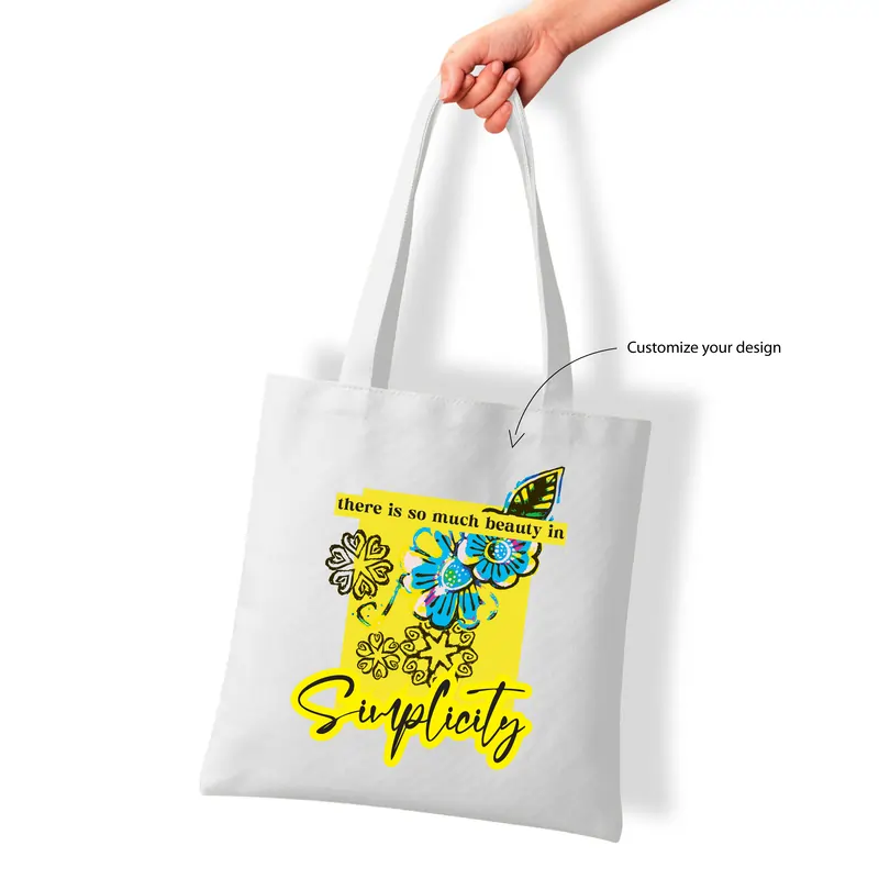 A hand holding a white tote bag featuring a yellow and blue floral design with the quote, "there is so much beauty in simplicity